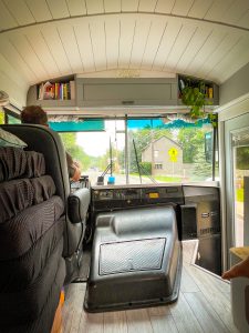 Photo of the interior drivers cabin and living room of the HildeBus while driving. Driver is sitting in the captains chair. There are bookshelves installed in the header. The black engine block is located in the center. The ceiling is white shiplap.