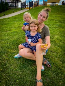 Mom and two daughters smiling and sitting in the grass. Mom has a glass of white wine in her hand.